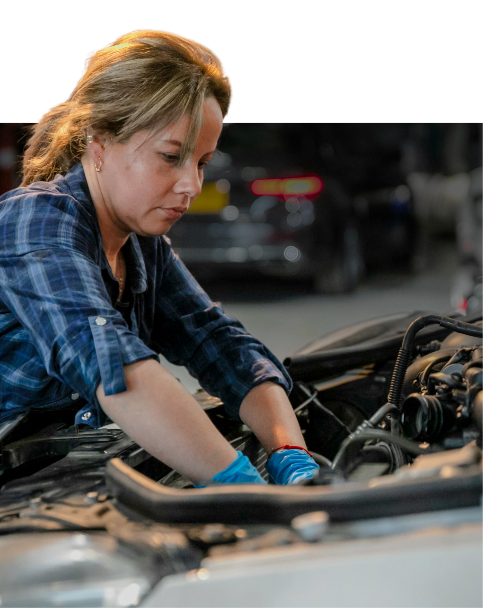 A woman in a plaid shirt and blue gloves works under the hood of a car in a garage, focusing intently on the engine.