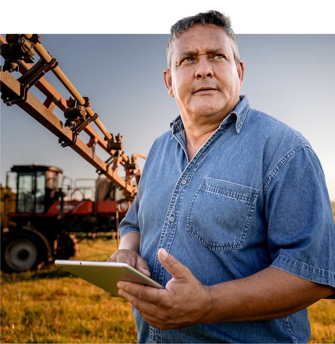 A middle-aged man in a denim shirt stands in a field holding a tablet, with agricultural machinery and clear blue sky in the background. He looks thoughtfully into the distance.