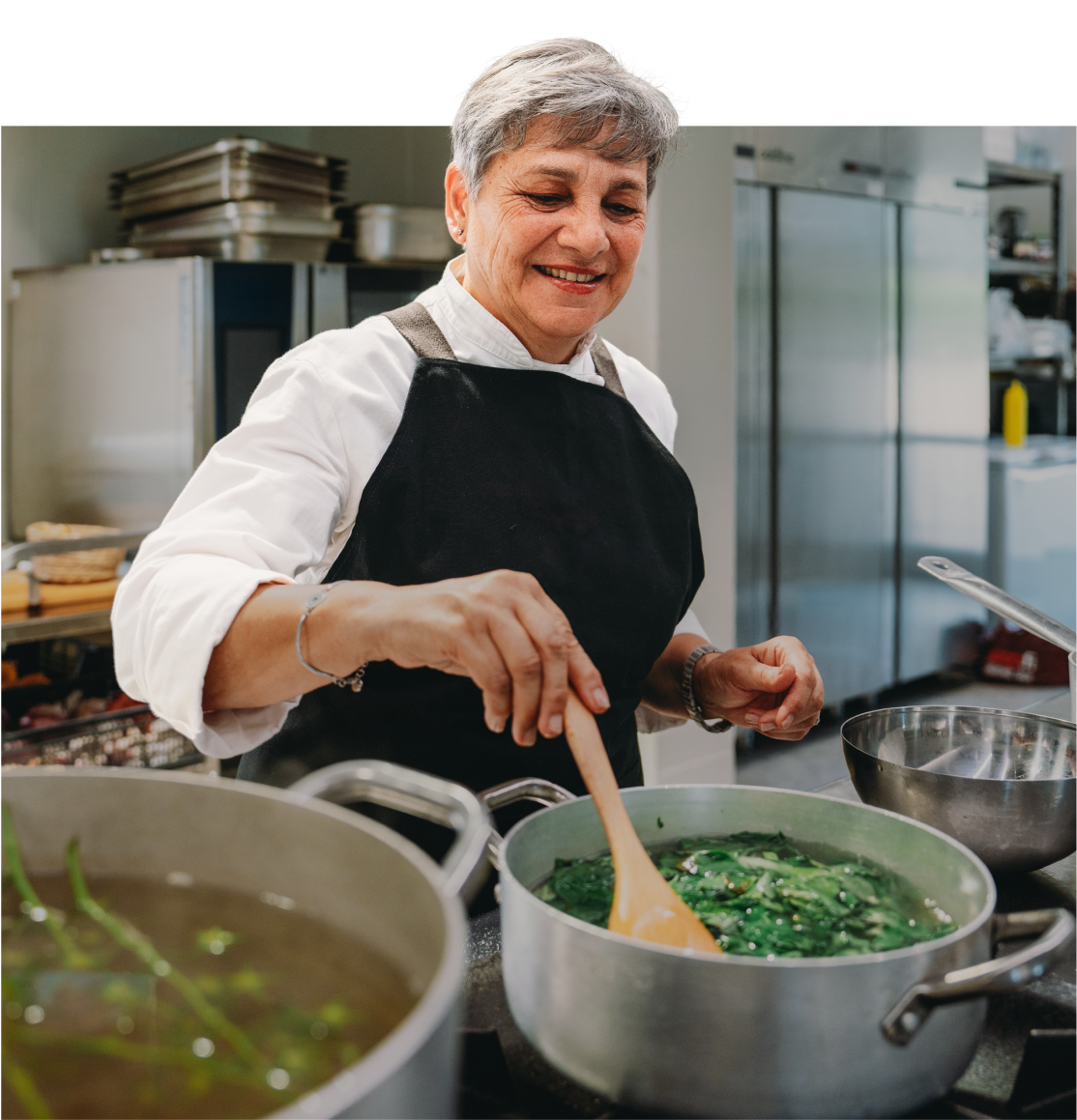 An older woman in a black apron stirs a pot of greens on a stovetop in a commercial kitchen, smiling as she cooks.