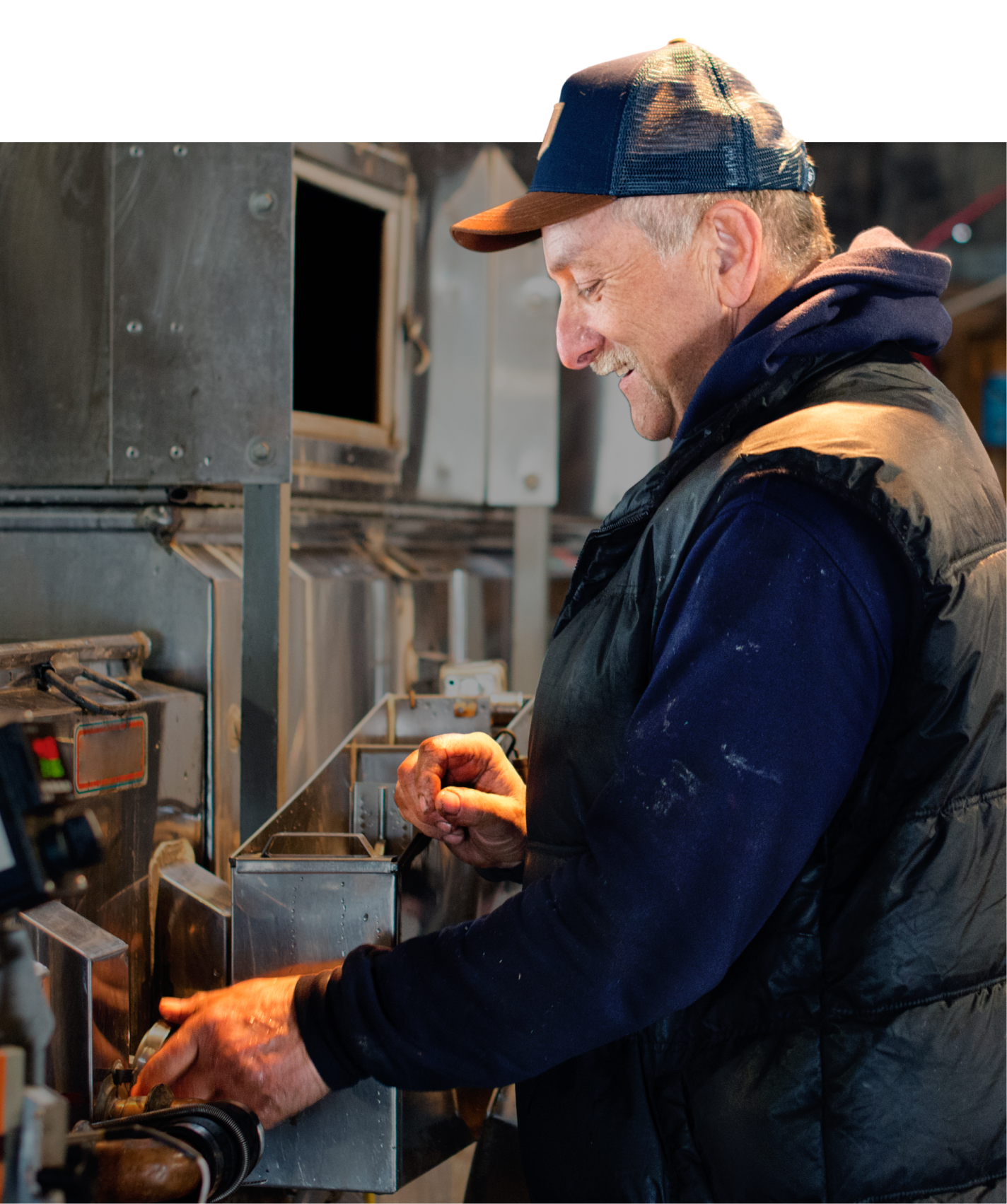 A man in a blue cap, navy hoodie, and black vest smiles while operating machinery in an industrial setting.