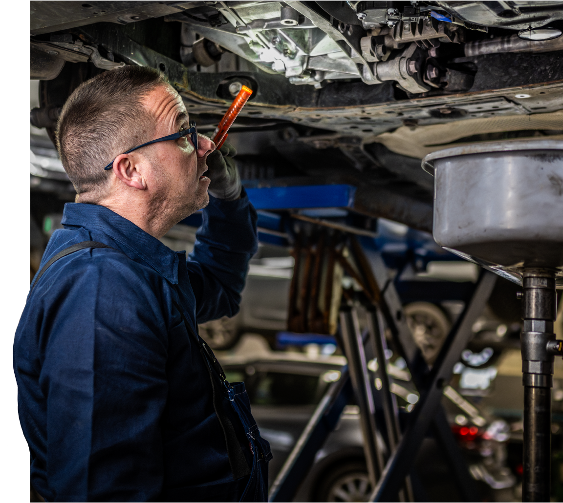 A mechanic wearing glasses and blue coveralls inspects the underside of a car with a flashlight in an auto repair shop, surrounded by car lifts and vehicles.